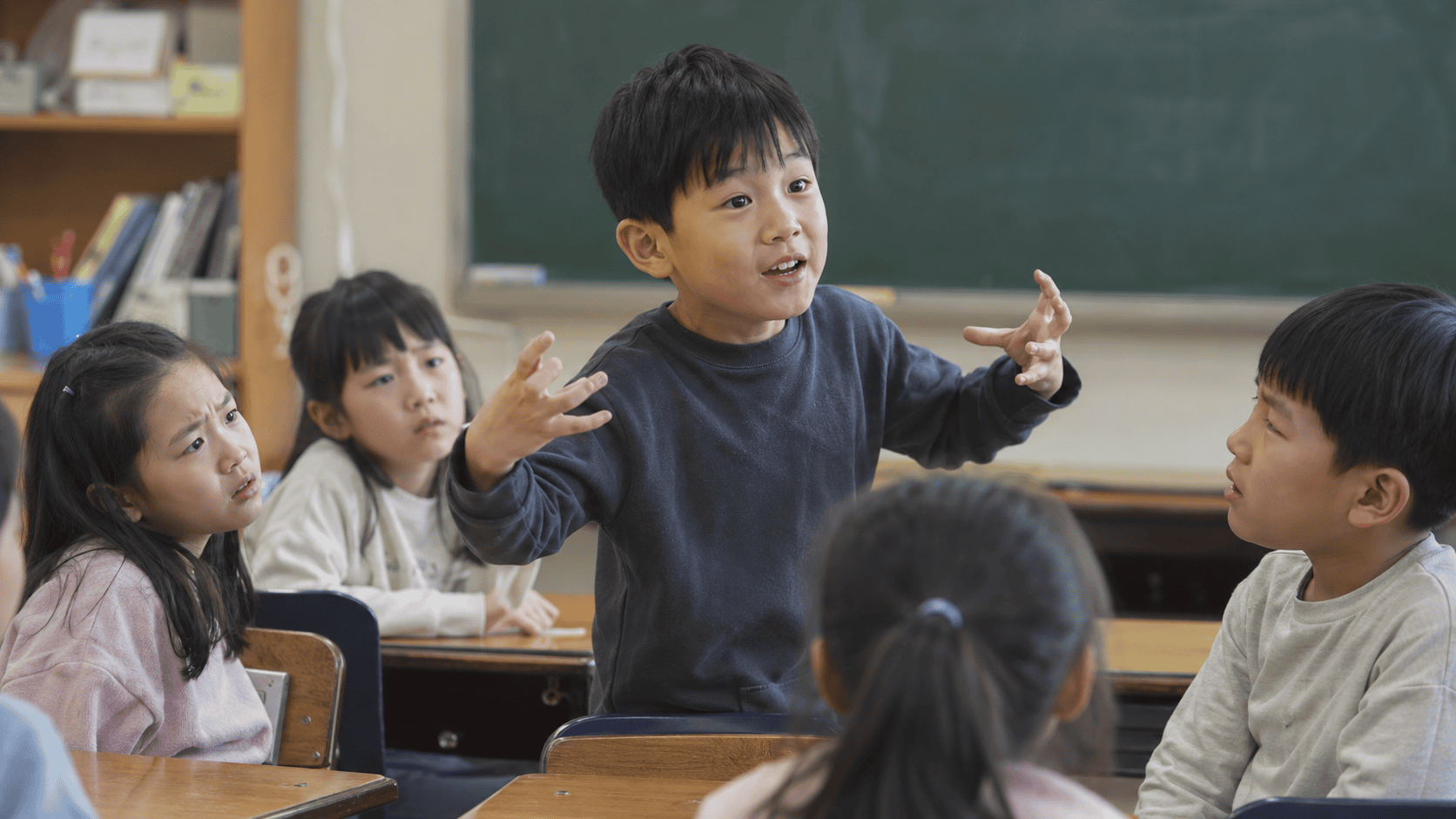 교실에서 남자아이가 손짓하며 발표하고 친구들이 경청한다.