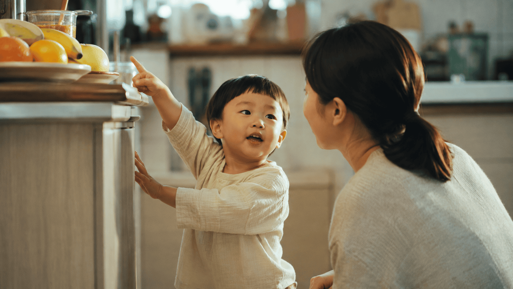 아이가 주방에서 엄마를 보며 손가락으로 과일을 가리킨다.