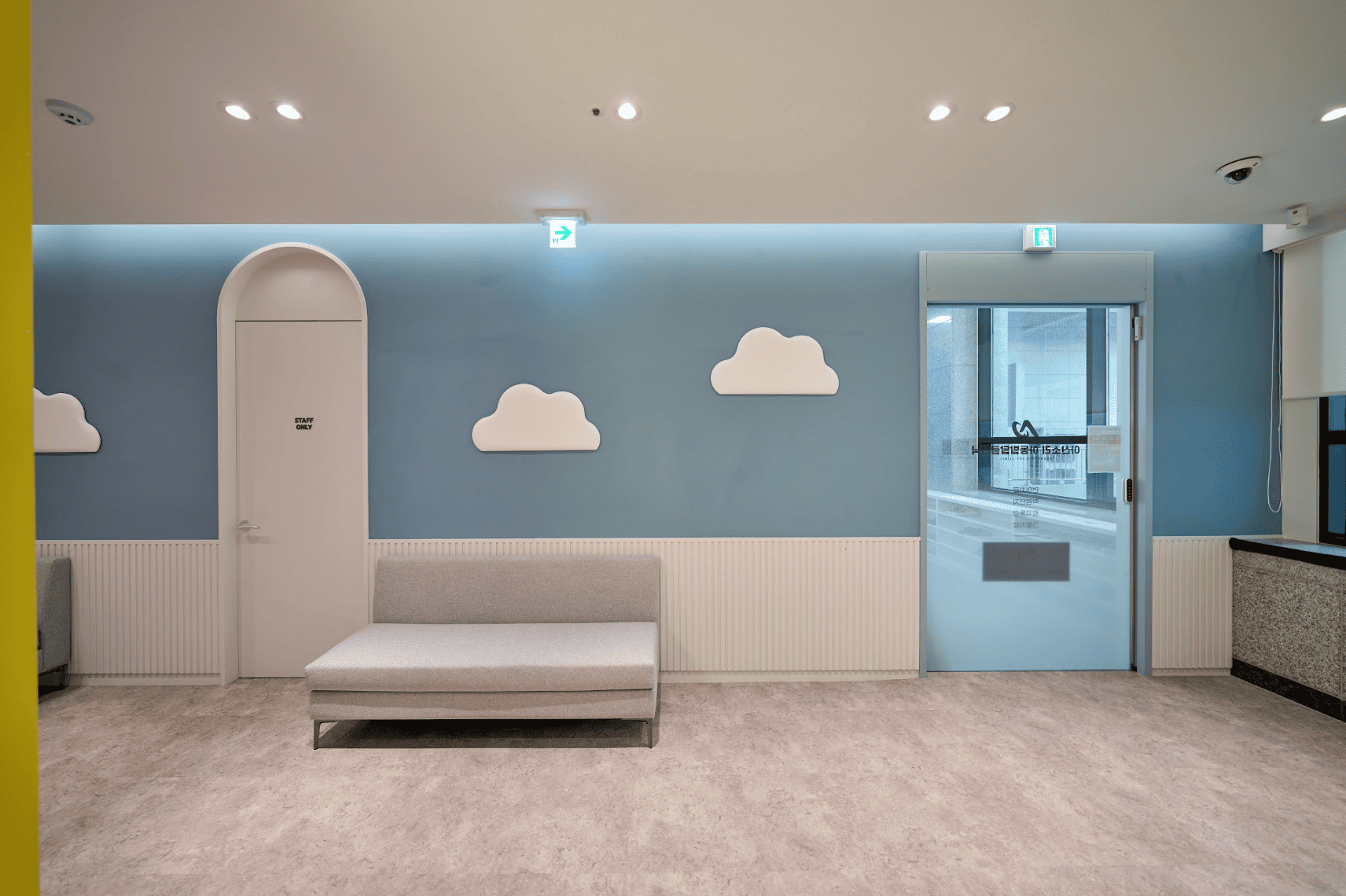 구름 장식이 있는 하늘색 벽과 회색 소파가 놓인 아동 치료실 복도.