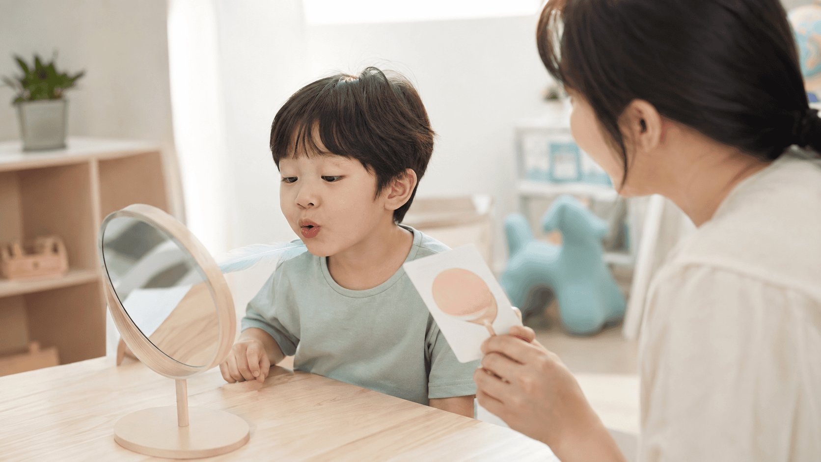 어린아이가 거울을 보며 깃털을 불고, 여성이 발음 교정 카드를 제시한다.